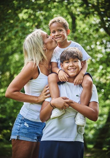 Femme embrassant un enfant, un autre enfant souriant sur son dos, entourés d'arbres.