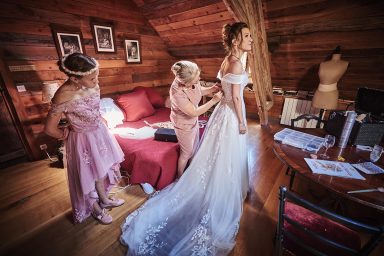 Femme en robe de mariée ajustée par deux accompagnatrices dans un cadre chaleureux.