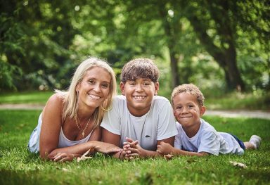 Une femme et deux enfants souriant, allongés sur l'herbe dans un parc verdoyant.