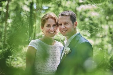 Un couple souriant en tenue de mariage, entouré de verdure.